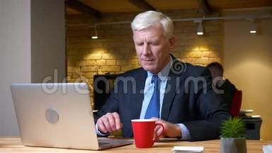 老白种人办公室工作人员使用笔记本电脑和在工作场所的办公室内喝咖啡的特写镜头